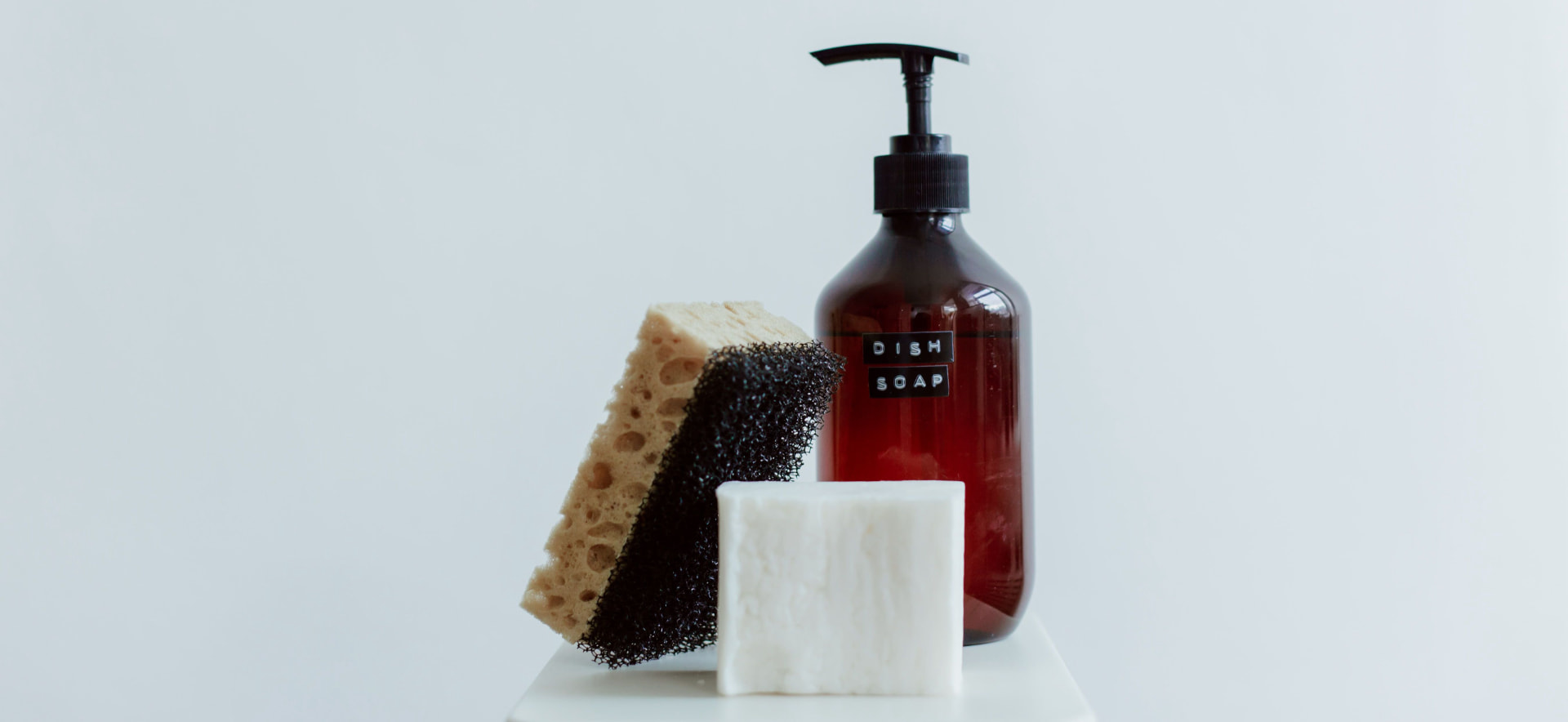 Amber dish soap dispenser with natural sponge, black scrubber and white soap bar on a clean counter